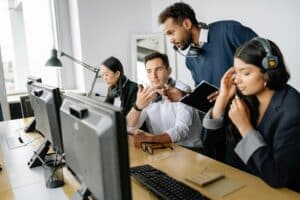 A computer trainer helping a client sat at a desk with 2 other individuals