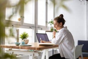 A woman in a white shirt sat in her work from home space with her laptop open whilst on her mobile, facing a window to a bright outdoor space.